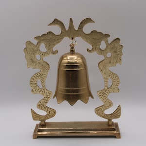 May include: A polished brass bell suspended from a decorative frame featuring two symmetrical dragon figures. The bell hangs from a central hook within the archway created by the dragons, all mounted on a rectangular brass base stand.
