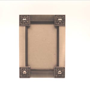 May include: A rectangular metal picture frame with a brushed finish. Each of the four corners features a square decorative accent containing a raised heart shape surrounded by a dotted pattern. The frame has a vertical orientation.