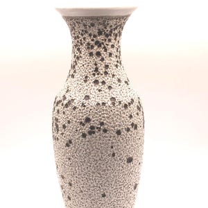 May include: A ceramic vase with a flared rim and a tapered neck, featuring a textured, speckled surface pattern of dark brown spots against a cream-colored base. The vessel has a rounded body that narrows toward the base and a smooth interior rim.