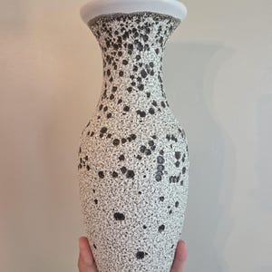 May include: A tall ceramic vase featuring a textured white base with a speckled pattern of dark brown and black spots. The vase has a flared rim, a narrow neck, and a rounded body that tapers towards the base, showcasing a distinct mottled finish.