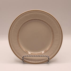 May include: A round cream-colored ceramic plate featuring a gold-toned Greek key pattern encircling the center. The rim is decorated with a textured, vertical ribbed design and a thin gold-colored edge, presented on a silver-toned wire stand.