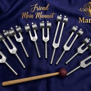 May include: A set of silver tuning forks with different frequencies, displayed on a dark blue cloth. A wooden mallet with a red head is placed near the forks. The image includes text that reads "Friend Mein Mannat" and "Mannat".