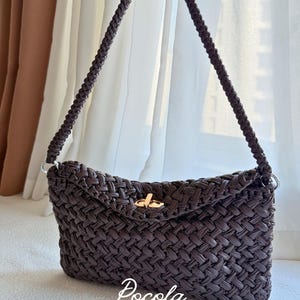 Peut inclure: Un sac à main tressé marron foncé avec une bandoulière tressée et un fermoir tournant en métal doré. L'avant du sac présente le texte blanc "Pocola ELEVATED DESIGN" imprimé près du bord inférieur de la matière tressée du sac à main.