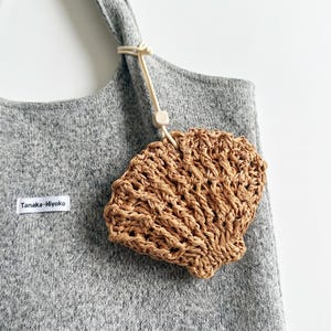 Puede incluir: Un bolso tote gris con un dije en forma de concha de rafia tejida colgando de la correa. El dije es de color marrón claro y está sujeto con un cordón color crema y una cuenta de madera. La etiqueta en la bolsa dice "Tanaka-Hiyoko."