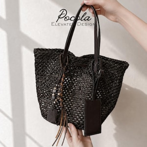 Può includere: Una borsa tote nera intrecciata con manico in pelle marrone scuro e targhetta per bagagli abbinata. La borsa è decorata con un ciondolo composto da cordini in pelle intrecciata e piccole perline. Il testo Pocola ELEVATE DESIGN è visibile.