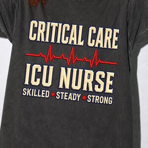 Puede incluir: Una camiseta gris carbón con texto bordado que dice CRITICAL CARE en color crema, una línea roja de ritmo cardíaco EKG, el texto ICU NURSE en crema y las palabras SKILLED, STEADY y STRONG separadas por pequeños puntos rojos en la parte inferior.
