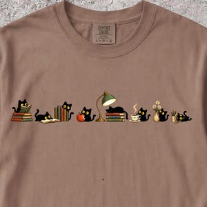 以下が含まれることがあります： ライトブラウンのTシャツ。本、緑色のデスクランプ、赤いリンゴ、湯気の立つコーヒーカップ、花瓶、鉛筆と戯れる数匹の小さな黒猫のグラフィックが描かれています。首元の内側のラベルには「Comfort Colors L-G-G」と記されています。