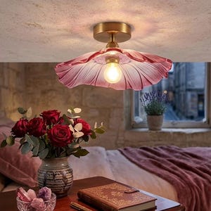 Puede incluir: Una lámpara de techo de cristal rosa con diseño de pétalos y base dorada. Un ramo de rosas rojas sobre una mesa de madera, junto a un libro y un cuenco de cristal. La escena crea un ambiente cálido y acogedor.