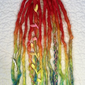 May include: A set of rainbow-colored dreadlocks, transitioning from red to yellow, green, and blue. The dreads are adorned with yellow and pink leaf accents and silver feather charms, creating a vibrant and bohemian aesthetic.