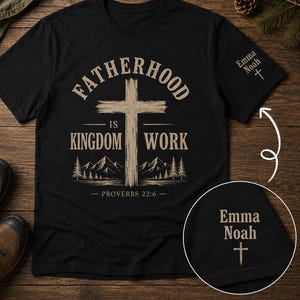 May include: Black t-shirt with the text "FATHERHOOD IS KINGDOM WORK" and a cross graphic. The shirt also features the name "Emma Noah" and a cross on the sleeve. The design includes mountain and tree illustrations.