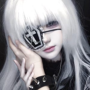 May include: A black and white eye patch featuring a coffin shape with a cross emblem and a silver chain detail. The eye patch is secured with a thin black strap and has a ruffled lace trim. It is worn with a black spiked wristband and long white hair.