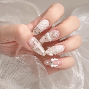 May include: Almond-shaped nails featuring a translucent base with white filigree patterns, silver-toned cross and heart charms, small star accents, a white bow, and a small pair of white wings with a central clear rhinestone embellishment.