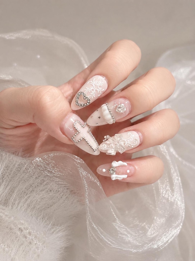 May include: Almond-shaped nails featuring a translucent base with white filigree patterns, silver-toned cross and heart charms, small star accents, a white bow, and a small pair of white wings with a central clear rhinestone embellishment.