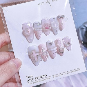 May include: A set of ten almond-shaped press-on nails in shades of pale pink and white, featuring 3D embellishments including a pink paw print, silver cross, heart charms, bows, stars, and rhinestones, displayed on a card labeled Nail ART STUDIO.