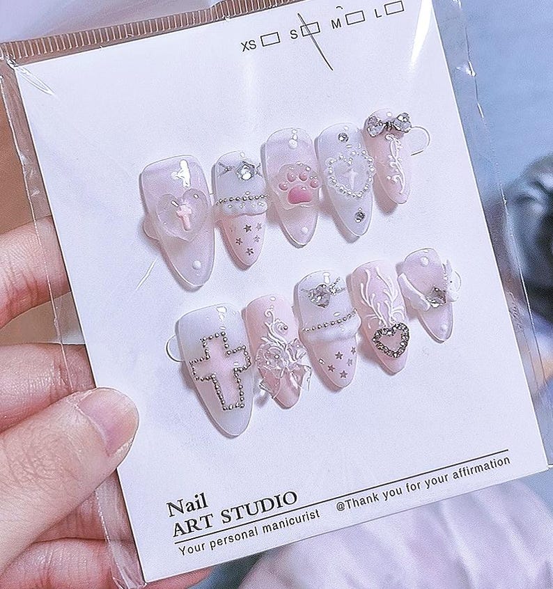 May include: A set of ten almond-shaped press-on nails in shades of pale pink and white, featuring 3D embellishments including a pink paw print, silver cross, heart charms, bows, stars, and rhinestones, displayed on a card labeled Nail ART STUDIO.