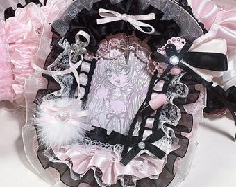 May include: A decorative oval frame featuring a central illustration of an anime character, surrounded by layers of black and pink ruffled fabric, white lace, black satin ribbons, a silver cross charm, a small white pom-pom, and a pink pill charm.