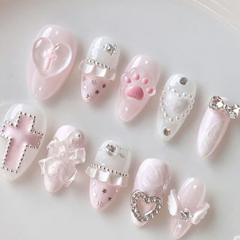 May include: A set of ten press-on nails in shades of pale pink and white. The designs feature 3D elements including a clear heart with a cross, a pink cat paw, silver stars, white ruffles, a pearl-bordered heart, a silver bow, and a heart charm.