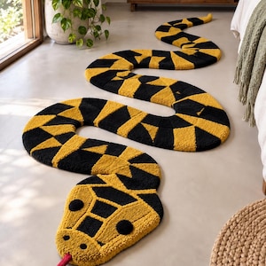 Puede incluir: Una alfombra larga y sinuosa con forma de serpiente que presenta un patrón geométrico repetitivo en amarillo mostaza y negro. La alfombra tiene una cabeza redondeada con dos ojos circulares negros, pequeñas fosas nasales y una lengua roja.