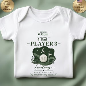 May include: A white baby bodysuit featuring a dark green graphic with the text PLAYER 1 Mom, PLAYER 2 Dad, and PLAYER 3. Below is a golf ball with baby footprints, a moon, stars, and the text Loading... ARRIVING 2027 Tiny Birdie. Big Dreams.