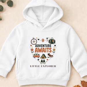 Peut inclure: Un sweat-shirt à capuche blanc avec un motif central en forme de cœur contenant le texte "ADVENTURE AWAITS" et "LITTLE EXPLORER". Le design inclut des illustrations d'une boussole, d'une valise, d'un van, d'une tente, d'une chaussure et de jumelles.