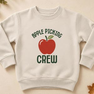 Könnte beinhalten: Ein beigefarbenes Langarm-Sweatshirt mit Rundhalsausschnitt und einer Grafik eines roten Apfels mit grünem Blatt. Der Text "APPLE PICKING" ist dunkelgrün über dem Apfel und das Wort "CREW" ist dunkelgrün unter dem Apfel aufgedruckt.