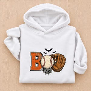 May include: A white hooded sweatshirt featuring a graphic print on the front. The design includes the orange letter B, a baseball, and a brown leather baseball glove. Black spiderwebs and three small black bats surround the central elements.