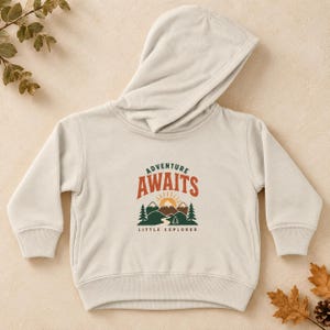 Peut inclure: Un sweat-shirt à capuche de couleur crème avec une impression graphique sur la poitrine. Le motif comprend le texte "ADVENTURE AWAITS" en orange et "LITTLE EXPLORER" en vert foncé, avec une illustration de montagnes, un soleil et des sapins.