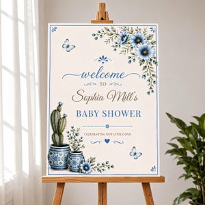 Puede incluir: Un letrero de bienvenida rectangular para el baby shower de Sophia Mill con ilustraciones florales azules, dos macetas con patrones azules y blancos con un cactus y flores, y dos mariposas sobre un fondo crema con bordes azules.