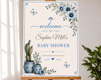 May include: A rectangular welcome sign for Sophia Mill's baby shower featuring blue floral illustrations, two blue and white patterned pots with a cactus and flowers, and two butterflies on a cream background with blue border lines and text.