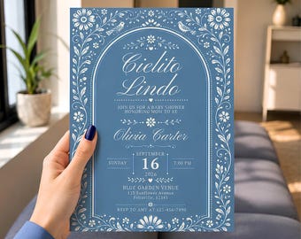 May include: A rectangular blue baby shower invitation card featuring white floral borders and an arched frame. The text reads Cielito Lindo, Join us for a baby shower honoring mom to be, Olivia Carter, September 16, 2026, 7:00 PM, Blue Garden Venue, 123 Sunflower Avenue, Felixville, 12345, RSVP to Amy at 123-456-7890.