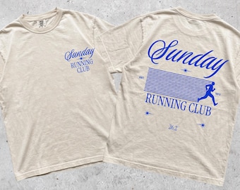 May include: A beige short-sleeve t-shirt featuring blue text and graphics. The front displays "Sunday RUNNING CLUB". The back features "Sunday RUNNING CLUB" in large script, a graphic of a runner, horizontal lines, "EST. 1972", and "26.2".