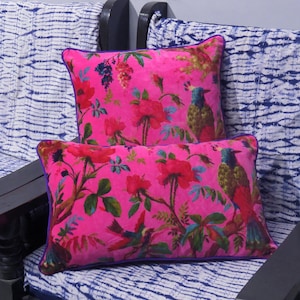 May include: Two rectangular throw pillows with a vibrant pink background. The pillows feature a floral and bird pattern in red, green, blue, and yellow. The pillows have a purple border and are placed on a blue and white patterned surface.