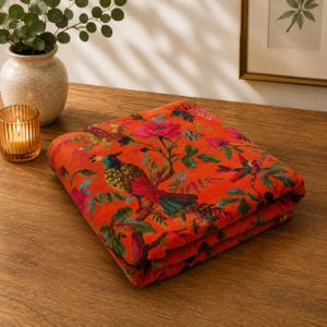 May include: Folded orange fabric with a vibrant floral and bird pattern. The design features a colorful bird perched on a branch with pink flowers and green leaves. The fabric is folded neatly on a wooden surface.