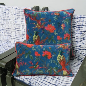 Könnte beinhalten: Zwei dekorative Kissen mit tealfarbenem Hintergrund und einem lebendigen Blumen- und Vogelmuster. Die Kissen haben eine rote Umrandung und sind auf einer gemusterten Oberfläche platziert. Das Design zeigt rote Blumen und bunte Vögel.