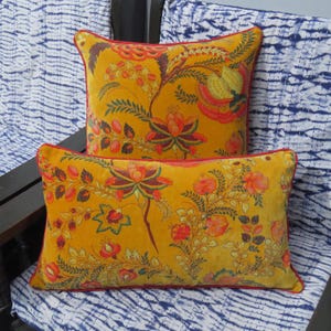 May include: Two decorative throw pillows in a vibrant mustard yellow velvet fabric, featuring a detailed floral pattern in shades of orange, red, and green. Each pillow is finished with a thin red piping along the edges for a defined border.