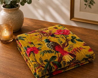 Puede incluir: Una manta de terciopelo color amarillo mostaza plegada con un estampado botánico vibrante de flores rosas y un pájaro multicolor con plumaje verde, marrón y rojo posado en una rama, que muestra una superficie de tela suave y texturizada.