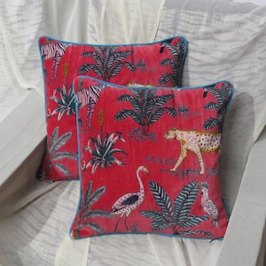 Könnte beinhalten: Zwei quadratische Kissen aus leuchtend rotem Samtstoff mit einem Druckmuster aus Zebras, Leoparden, Flamingos und tropischen Pflanzen. Jedes Kissen ist mit einer leuchtend blauen Paspelierung versehen, die einen deutlichen Kontrast bildet.