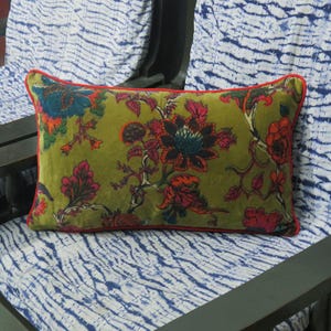 Peut inclure: Un coussin rectangulaire en velours vert olive avec un motif floral rouge, orange et turquoise. Le coussin a une bordure rouge et est posé sur une surface à motifs bleus et blancs. Dimensions : environ 40 cm x 60 cm.