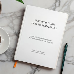 Puede incluir: Un libro blanco titulado "PRACTICAL GUIDE HOW TO READ LABELS" con texto sobre cómo evitar errores de compra. Se ve un marcador naranja y un bolígrafo estampado. El subtítulo del libro menciona Gluten, Lactosa, Fructosa, Polioles, Histamina y FODMAP.
