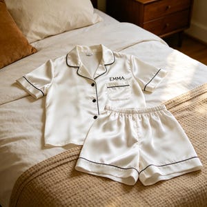 May include: Ivory satin pajama set with black trim. The short-sleeved top has a breast pocket and the name "EMMA" embroidered on it. The matching shorts have an elastic waistband. The set is displayed on a bed with a neutral-toned blanket.