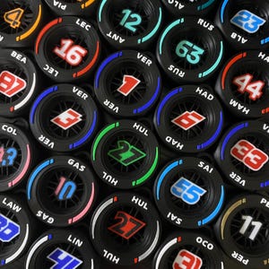 Puede incluir: Una colección de objetos circulares negros que se asemejan a neumáticos, cada uno con un número diferente e iniciales de piloto en varios colores. Los objetos tienen un diseño central y un borde con texto. Los números son en blanco, rojo, azul y verde.