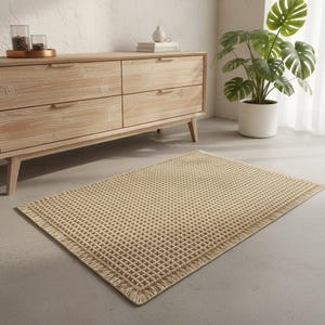 Op de afbeelding: Een rechthoekig beige tapijt met een wafelpatroon en gefranjerde randen. Het tapijt ligt op een grijze vloer voor een licht houten dressoir en een potplant.