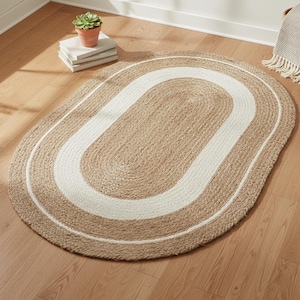 May include: Oval jute rug with a natural tan color and a white border. The rug is made of woven jute fibers and is placed on a hardwood floor. The rug's design is simple and neutral.