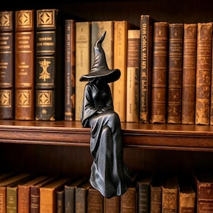 May include: A matte black figurine of a seated figure wearing a wide-brimmed pointed hat and a long, flowing robe. The figure is positioned with its legs dangling over the edge of a shelf, featuring a smooth, dark finish and a draped garment.