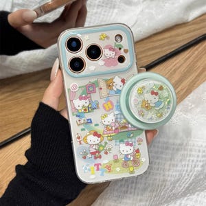 May include: Clear phone case with a light blue border and a matching pop socket. The case features a colorful Hello Kitty design with various scenes and characters. The pop socket has a glittery Hello Kitty design.