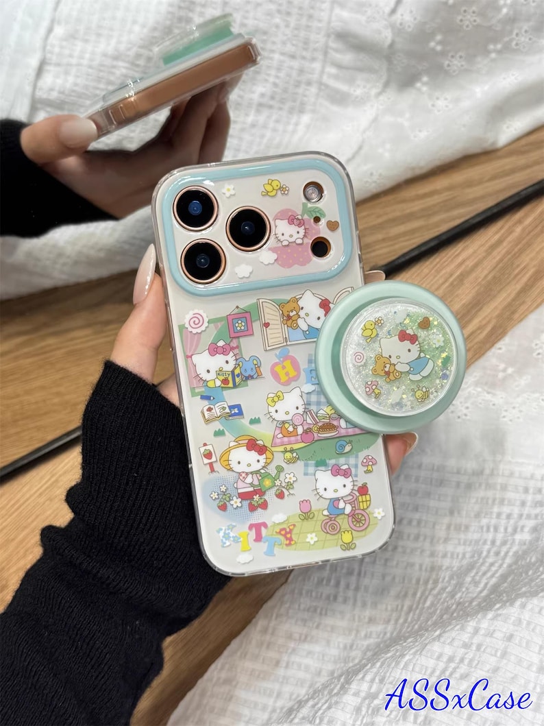 May include: Clear phone case with a light blue border and a matching pop socket. The case features a colorful Hello Kitty design with various scenes and characters. The pop socket has a glittery Hello Kitty design.