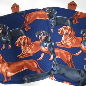 May include: Two blue pot holders with a pattern of dachshund dogs in black, brown, and tan. The dogs are in various poses, including standing, sitting, and lying down.