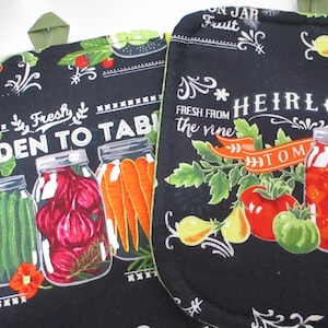 May include: Black oven mitts with a garden theme. One mitt features jars of vegetables and the text "GARDEN TO TABLE." The other mitt has tomatoes and the text "HEIRLOOM TOM." Both mitts have a black background with white and orange accents.