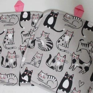 May include: A pair of gray oven mitts with a pink hanging loop. The mitts are covered in a repeating pattern of black and white cats.