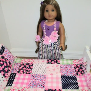 May include: A doll stands in a white bed with a patchwork quilt and pillow. The quilt features pink, white, and navy floral and polka dot patterns. The doll wears a purple dress with a floral necklace and bracelet.